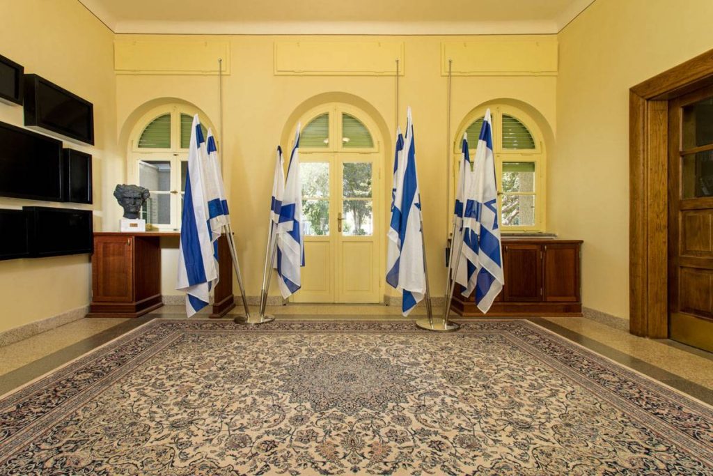 צילום עיצוב פנים של לובי היסטורי בבית בן־גוריון עם חלונות קשת, ריצוף מקורי ודגלי ישראל.